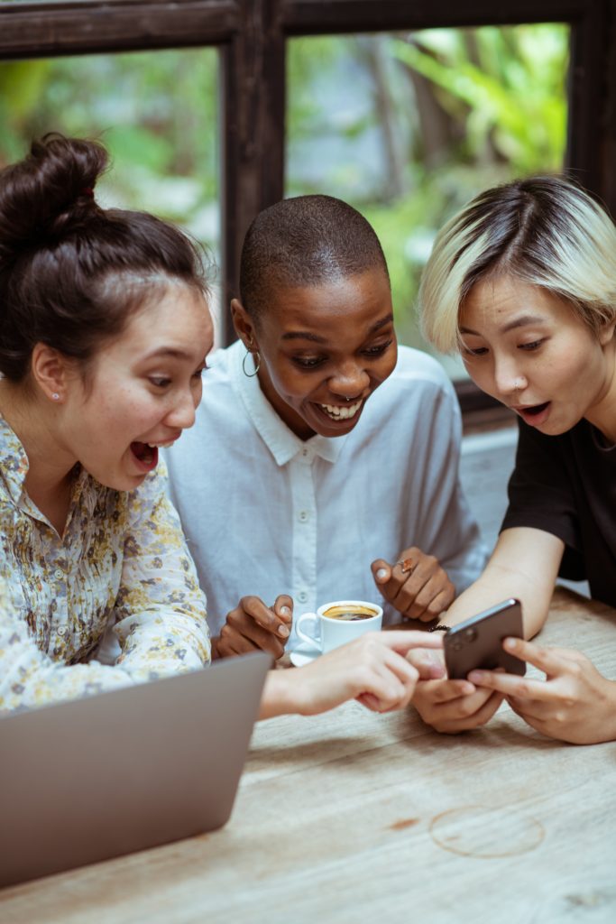 6 Ways To Promote Events On Social Media 4 Amazed Surprized Multiethnic Female Friends In Casual Clothes With Cup Of Coffee Browsing Smartphone While Sitting At Wooden Table With Laptop Against Window
